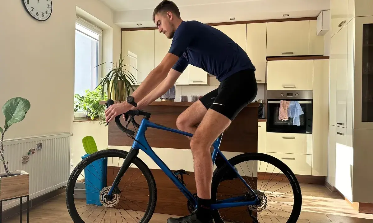 Mężczyzna w pozycji na rowerze gravel, trenuje w domu. W tle kuchnia i zegar.