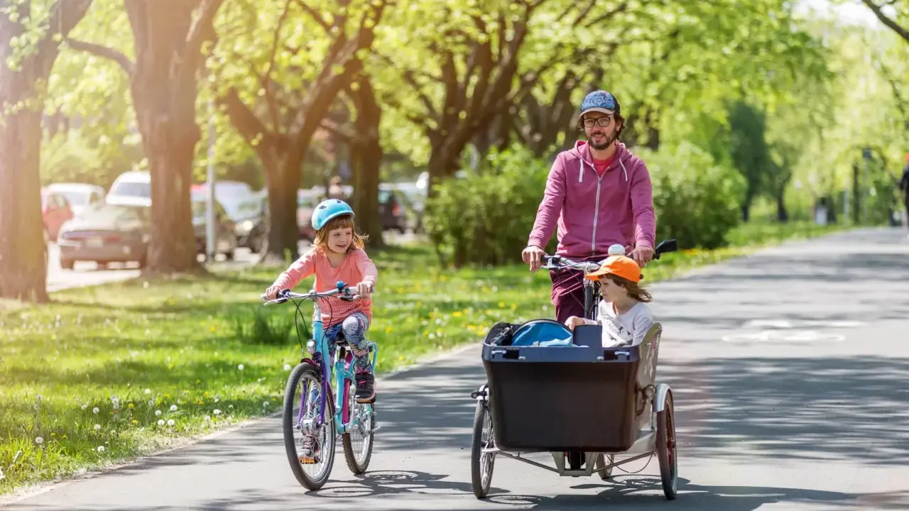 Radosne dziecko na rowerze jedzie przed rodzicem, który wiezie drugie dziecko w przyczepce rowerowej.