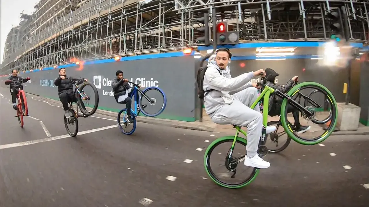 Młodzi mężczyźni wykonują **jazdę na jednym kole** na rowerach, mijając plac budowy. Jeden z nich, w szarym dresie, robi to na zielonym rowerze.