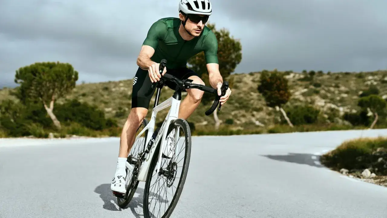 Kolarz w zielonej koszulce i białym kasku w aerodynamicznej pozycji na rowerze szosowym, pokonujący krętą drogę wśród pagórków.