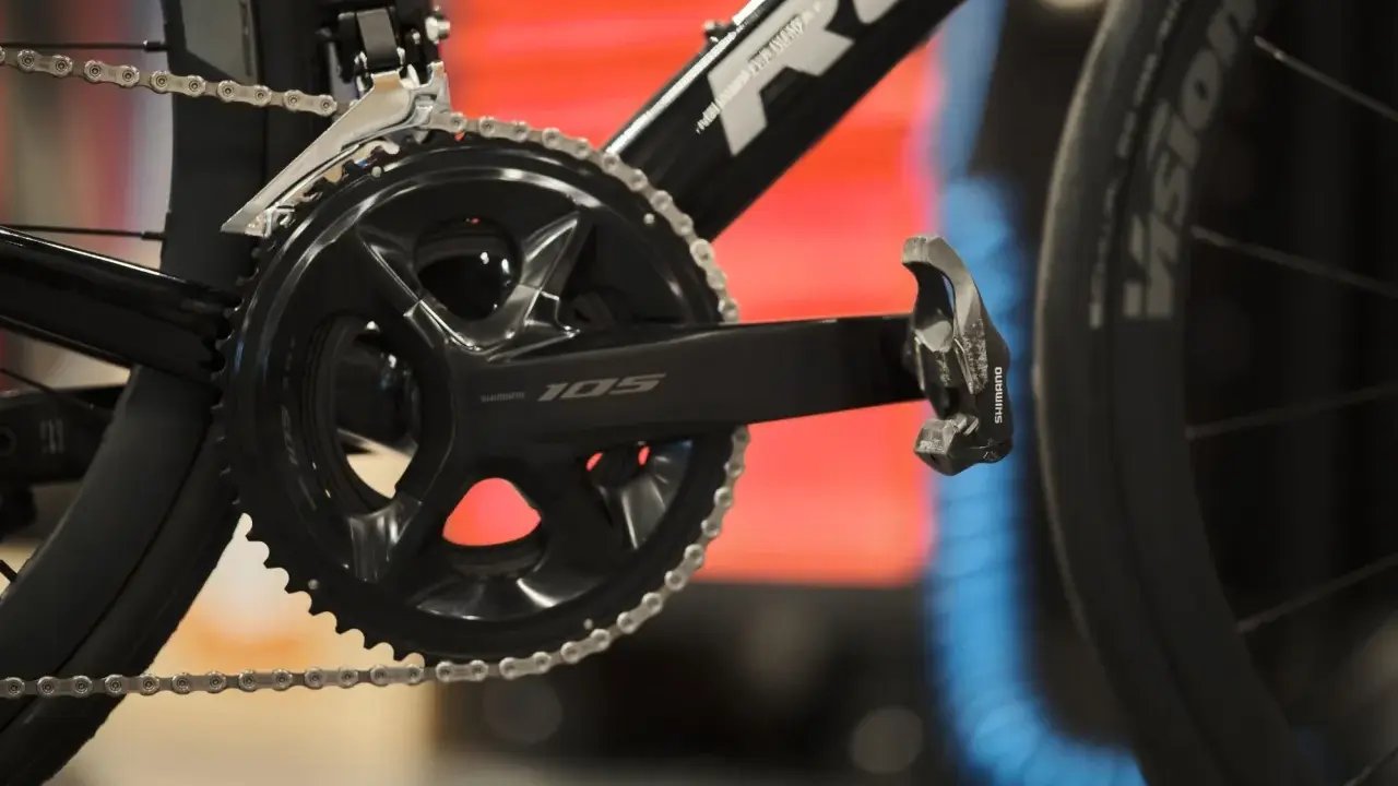 Szukasz informacji, jak dobrać korbę do roweru? Zobacz zbliżenie na czarną korbę Shimano 105 z łańcuchem i pedałem.