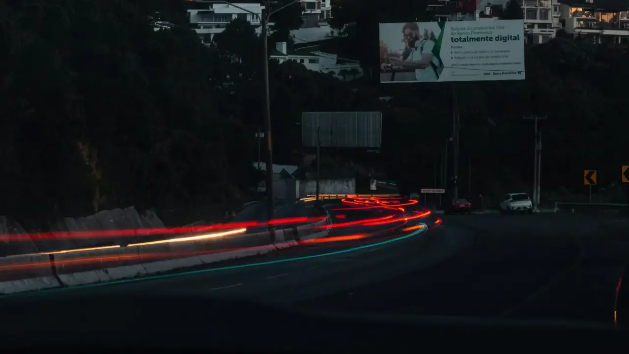 Smugi świateł samochodowych na drodze nocą, sugerujące średnią prędkość. W tle widać domy na wzgórzu i billboard.