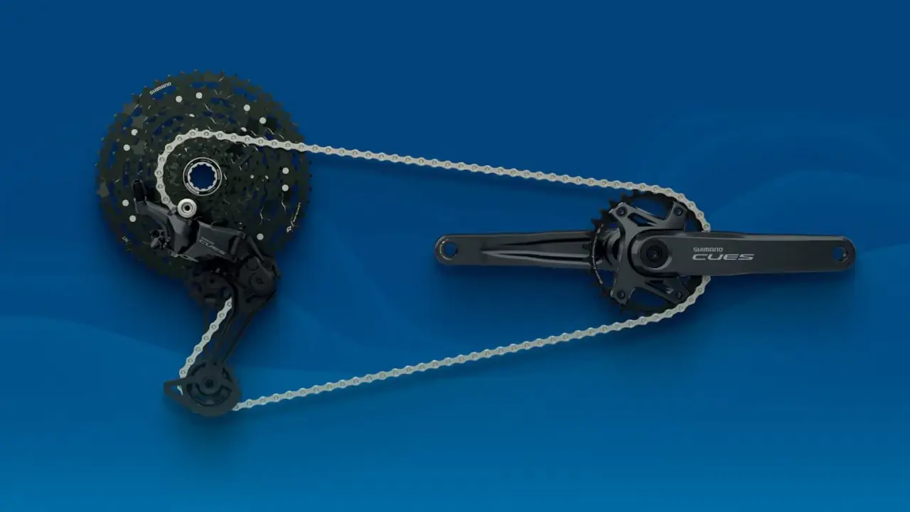 Zestaw napędowy Shimano CUES: korba, przerzutka i kaseta z łańcuchem na niebieskim tle.