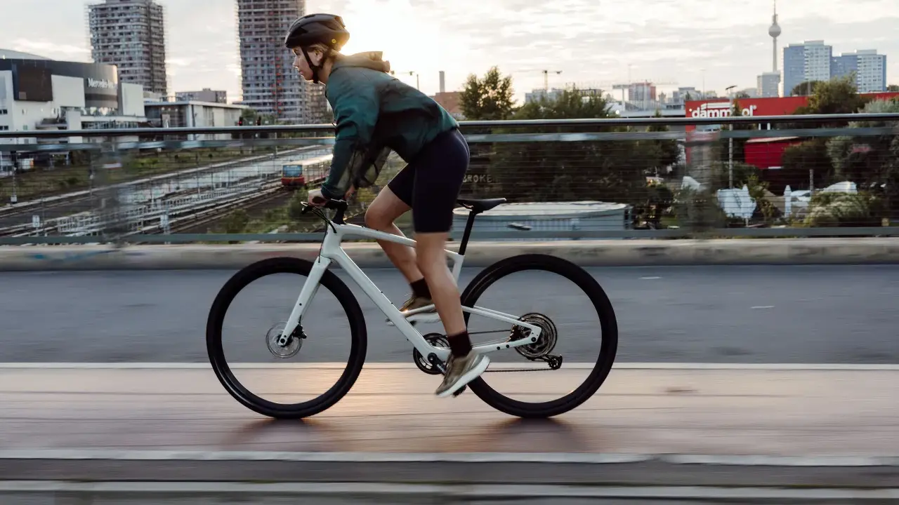Kobieta w kasku i sportowym stroju jedzie na białym rowerze crossowym po moście, z miejskim krajobrazem w tle.