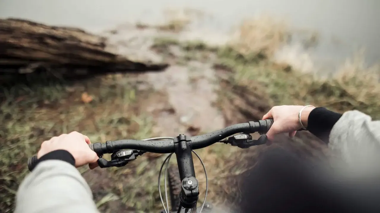 Ręce na kierownicy roweru, błotnista ścieżka i widok na wodę. To właśnie dlatego warto jeździć rowerem – bliskość natury i wolność.