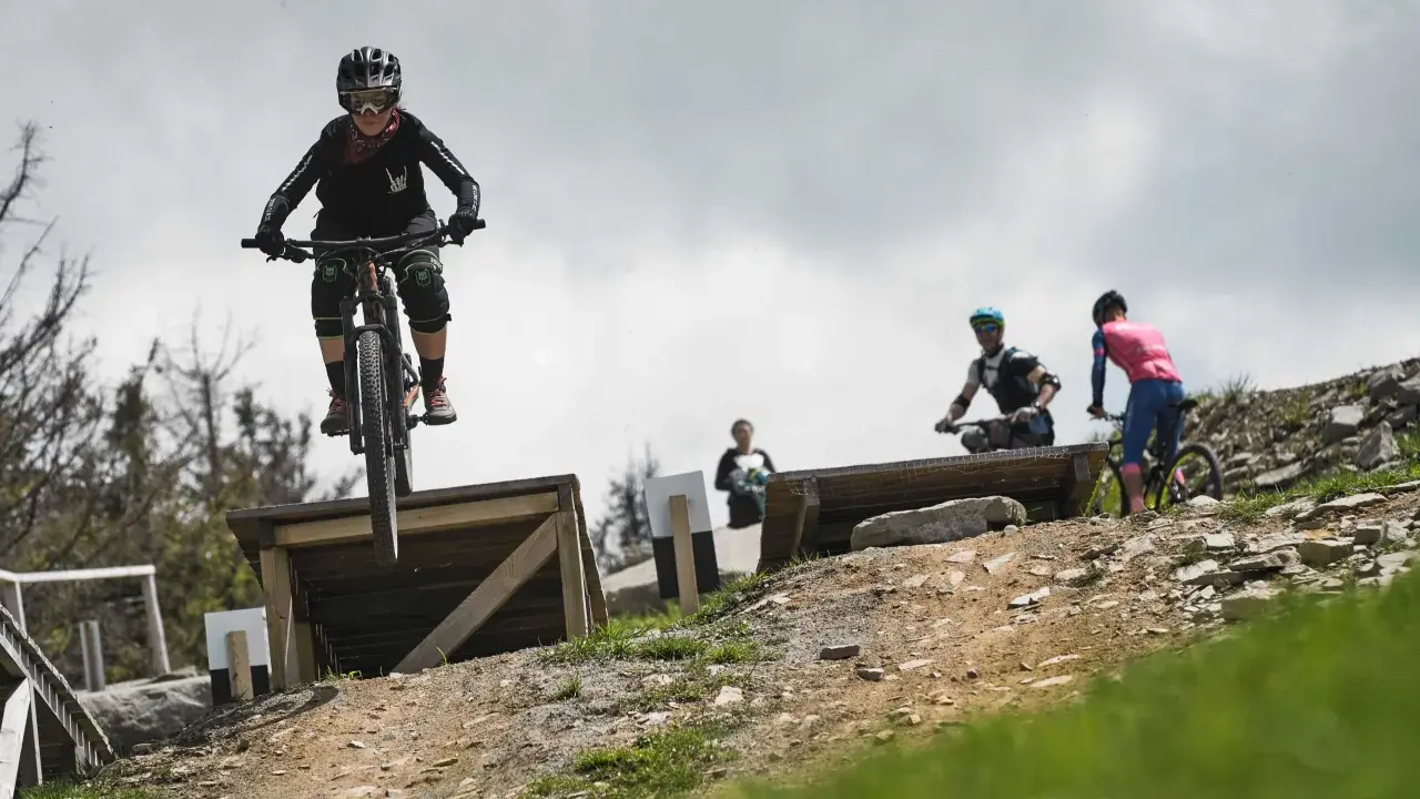 Młoda rowerzystka w kasku i goglach skacze na rowerze w Szczyrk Bike Park. W tle inni rowerzyści.
