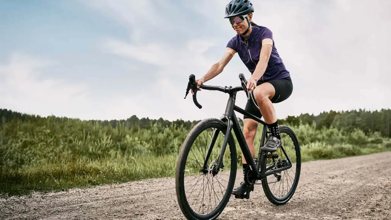 Kobieta na rowerze **gravel** pokonuje szutrową drogę. W tle las i niebo.