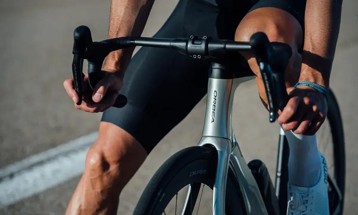 Kolarz w czarnych spodenkach i białych skarpetkach trzyma kierownicę aerodynamicznego roweru Orbea. Szytka rowerowa jest gotowa do jazdy.