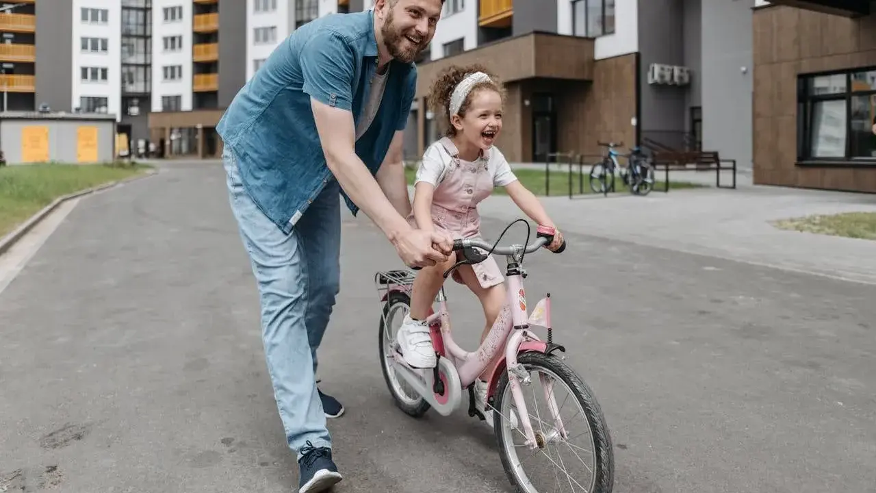 Tata uczy córkę, jak nauczyć dziecko jazdy na rowerze. Radosna dziewczynka na różowym rowerze, tata ją asekuruje.