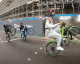 Młodzi mężczyźni wykonują **jazdę na jednym kole** na rowerach, mijając plac budowy. Jeden z nich, w szarym dresie, robi to na zielonym rowerze.