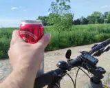 Rowerzysta trzyma puszkę napoju, co sugeruje kierowanie rowerem w stanie nietrzeźwości. Na kierownicy widoczny licznik.