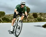 Kolarz w zielonej koszulce i białym kasku w aerodynamicznej pozycji na rowerze szosowym, pokonujący krętą drogę wśród pagórków.