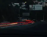 Smugi świateł samochodowych na drodze nocą, sugerujące średnią prędkość. W tle widać domy na wzgórzu i billboard.