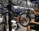 W warsztacie fabryki rowerów mechanik naprawia rower. W tle wiszą rowery i narzędzia.
