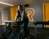 Uśmiechnięta kobieta na rowerze stacjonarnym, gotowa do codziennej jazdy na rowerze. Za nią neonowy napis "R".