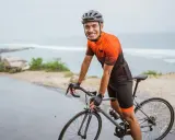 Uśmiechnięty kolarz w kasku i stroju sportowym na tle morza. Zastanawiasz się mips co to? To technologia w kasku, która zwiększa bezpieczeństwo.