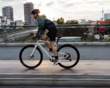 Kobieta w kasku i sportowym stroju jedzie na białym rowerze crossowym po moście, z miejskim krajobrazem w tle.