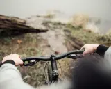 Ręce na kierownicy roweru, błotnista ścieżka i widok na wodę. To właśnie dlatego warto jeździć rowerem – bliskość natury i wolność.