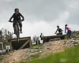Młoda rowerzystka w kasku i goglach skacze na rowerze w Szczyrk Bike Park. W tle inni rowerzyści.
