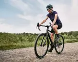 Kobieta na rowerze **gravel** pokonuje szutrową drogę. W tle las i niebo.