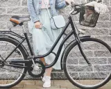Elegancka kobieta w zwiewnej sukience i dżinsowej kurtce opiera się o rower. Widoczny rozmiar ramy roweru sugeruje, że jest to model miejski, idealny na przejażdżki po mieście.