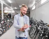 Uśmiechnięty mężczyzna notuje w zeszycie, otoczony rzędami rowerów różnych marek w magazynie.