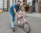 Tata uczy córkę, jak nauczyć dziecko jazdy na rowerze. Radosna dziewczynka na różowym rowerze, tata ją asekuruje.