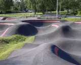Asfaltowy pump track z czerwonymi pasami, otoczony zielenią i drzewami. Idealne miejsce do zabawy na rowerze lub hulajnodze.