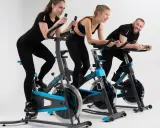 Trzy osoby, dwie kobiety i mężczyzna, ćwiczą na rowerach stacjonarnych. Wszyscy uśmiechnięci, widać, że lubią spinning.