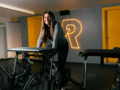 Uśmiechnięta kobieta na rowerze stacjonarnym, gotowa do codziennej jazdy na rowerze. Za nią neonowy napis "R".
