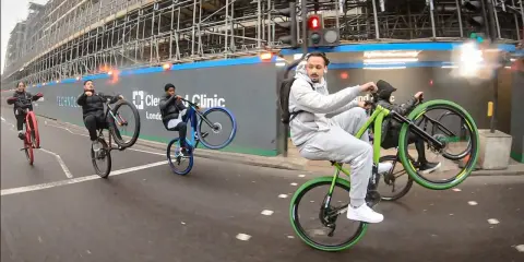 Młodzi mężczyźni wykonują **jazdę na jednym kole** na rowerach, mijając plac budowy. Jeden z nich, w szarym dresie, robi to na zielonym rowerze.