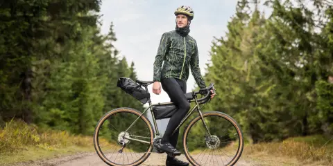 Wysoki mężczyzna w kasku i kurtce rowerowej pozuje na tle lasu, opierając się o swój gravelowy rower. Idealny rower dla wysokiego mężczyzny na leśne wyprawy.