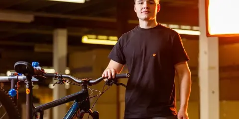 Młody mężczyzna w czarnej koszulce z logo R stoi obok roweru górskiego. To idealne miejsce, by znaleźć nowe portale rowerowe.