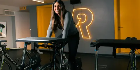Uśmiechnięta kobieta na rowerze stacjonarnym, gotowa do codziennej jazdy na rowerze. Za nią neonowy napis "R".