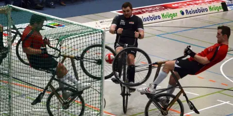 Gracze w piłkę rowerową walczą o piłkę przy bramce. Jeden z zawodników próbuje strzelić gola.