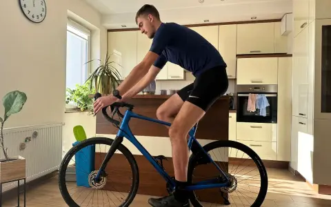 Mężczyzna w pozycji na rowerze gravel, trenuje w domu. W tle kuchnia i zegar.