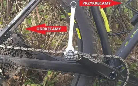 Klucz do pedałów rowerowych przy korbie, z czerwonymi strzałkami wskazującymi "Odkręcamy" i "Przykręcamy".