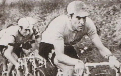 Dwóch kolarzy w akcji. Na pierwszym planie widać zawodnika w czarnych spodenkach i jasnej koszulce, zdeterminowanego jak zawsze Eddy Merckx.