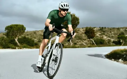 Kolarz w zielonej koszulce i białym kasku w aerodynamicznej pozycji na rowerze szosowym, pokonujący krętą drogę wśród pagórków.