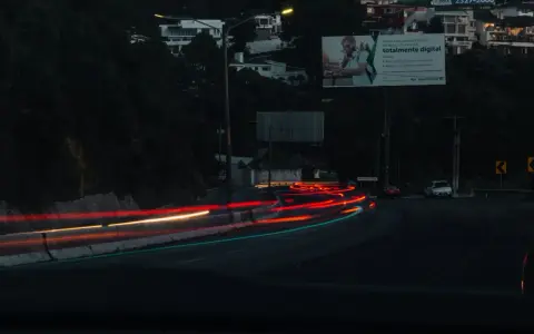 Smugi świateł samochodowych na drodze nocą, sugerujące średnią prędkość. W tle widać domy na wzgórzu i billboard.