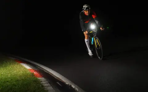 Kolarz w kasku z czaszką jedzie nocą, oświetlając drogę mocnym światłem. Czy rower musi mieć włączone światła w dzień? To pytanie nabiera sensu, gdy widzimy taką jazdę.