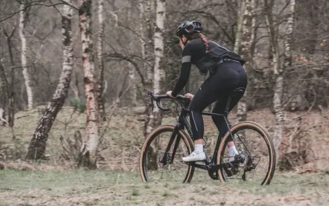 Kobieta w kasku i czarnym stroju jedzie na rowerze typu gravel przez las.