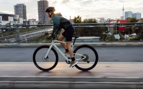 Kobieta w kasku i sportowym stroju jedzie na białym rowerze crossowym po moście, z miejskim krajobrazem w tle.