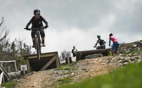 Młoda rowerzystka w kasku i goglach skacze na rowerze w Szczyrk Bike Park. W tle inni rowerzyści.
