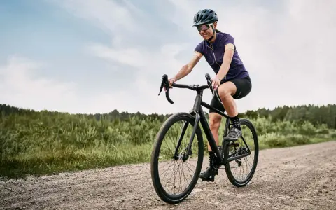 Kobieta na rowerze **gravel** pokonuje szutrową drogę. W tle las i niebo.
