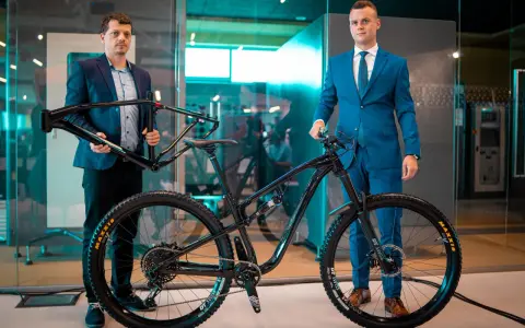 Dwóch mężczyzn prezentuje nowy rower full-suspension. Jeden trzyma ramę, drugi stoi przy gotowym rowerze. To duma firmy Romet.