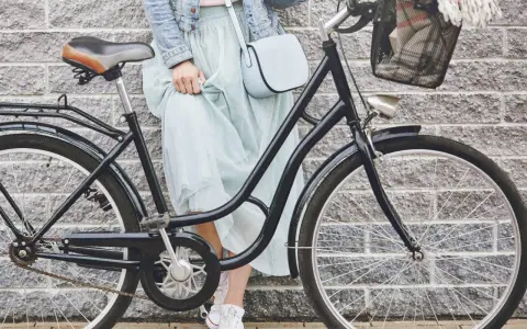 Elegancka kobieta w zwiewnej sukience i dżinsowej kurtce opiera się o rower. Widoczny rozmiar ramy roweru sugeruje, że jest to model miejski, idealny na przejażdżki po mieście.