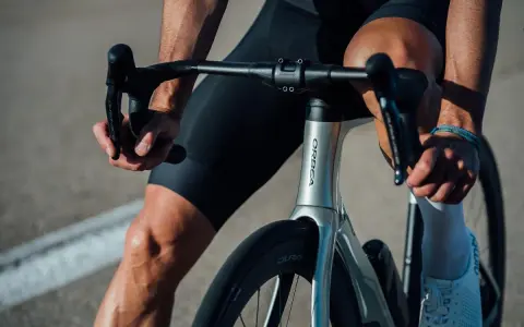 Kolarz w czarnych spodenkach i białych skarpetkach trzyma kierownicę aerodynamicznego roweru Orbea. Szytka rowerowa jest gotowa do jazdy.