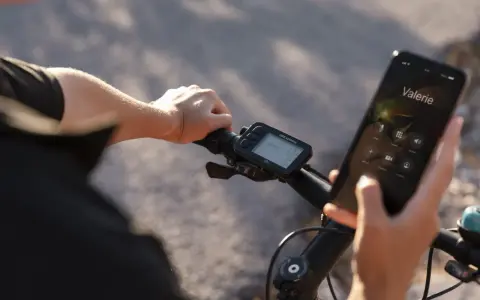 Rowerzysta trzyma telefon, na którym wyświetla się połączenie z Valerie. Obok widoczne są symbole na liczniku rowerowym, pokazujące prędkość i inne dane.
