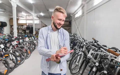 Uśmiechnięty mężczyzna notuje w zeszycie, otoczony rzędami rowerów różnych marek w magazynie.